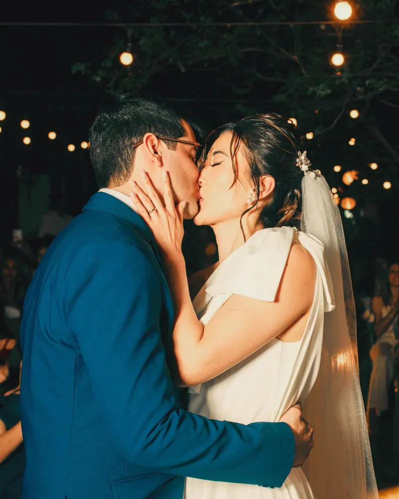 A couple kiss under string lights while their guests clap